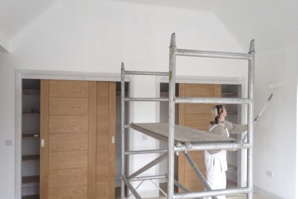 woman painting walls white with paint roller, scaffolding in the middle of the picture